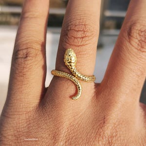 May include: A gold-colored snake ring. The ring wraps around the finger, with the snake's head at the top. The snake's body has a textured, scale-like pattern. The ring is a piece of jewelry.