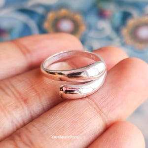 Könnte beinhalten: Ein polierter Ring in Silberoptik mit offenem Design und abgerundeten, zulaufenden Enden. Die Metalloberfläche ist stark reflektierend und zeigt eine klare, durchgehende Kurve, die sich teilweise um den Finger legt.