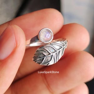 May include: A silver ring with a moonstone and leaf design. The ring features a round, iridescent moonstone set on a thin band. The band wraps around the finger with a detailed leaf design. The text "LuxeSparkStone" is visible.