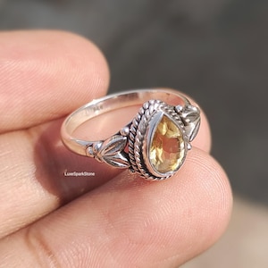 Könnte beinhalten: Ein silberner Ring mit einem birnenförmigen, gelben Edelstein. Der Ring hat aufwendige Details um den Stein und am Band, einschließlich Blatt- und Seilmuster. Das LuxeSparkStone-Logo ist sichtbar.