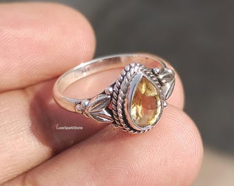 May include: A silver ring with a pear-shaped, yellow gemstone. The ring features intricate detailing around the stone and on the band, including leaf and rope-like designs. The ring is held in a hand, with the LuxeSparkStone logo visible.