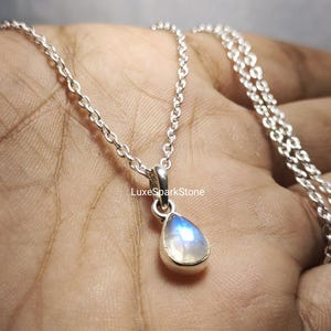 Puede incluir: Un collar de plata con un colgante de piedra lunar iridiscente en forma de pera. El colgante está engastado en un bisel de plata y cuelga de una fina cadena. El texto "LuxeSparkStone" es visible.