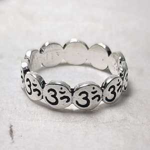 May include: A silver ring featuring a repeating pattern of the Om symbol. The ring has a circular design with the Om symbol in black on each of the ring's rounded sections. The ring is shown on a white surface.
