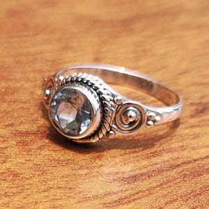 Könnte beinhalten: Ein silberner Ring mit einem großen, runden, hellblauen Edelstein. Der Ring hat ein dekoratives Band mit einem geflochtenen Design um den Edelstein und spiralförmige Details an den Seiten. Der Ring liegt auf einer Holzoberfläche.