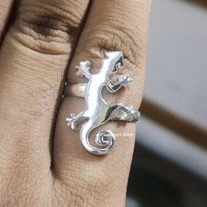 May include: A silver-colored ring featuring a lizard design. The lizard is wrapped around the finger, with its tail forming a spiral. The ring is made of a shiny metal and is shown on a finger.