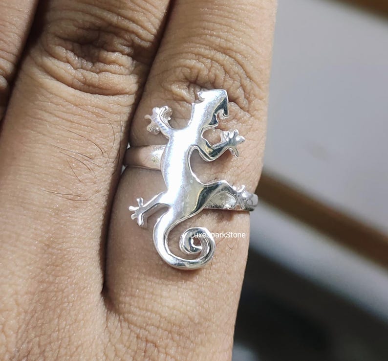 May include: A silver-colored ring featuring a lizard design. The lizard is wrapped around the finger, with its tail forming a spiral. The ring is made of a shiny metal and is shown on a finger.