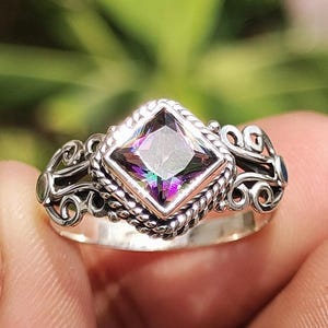 May include: A silver ring with a square, iridescent gemstone. The gemstone is set in a decorative bezel with a twisted rope design. The ring features ornate scrollwork on the sides.
