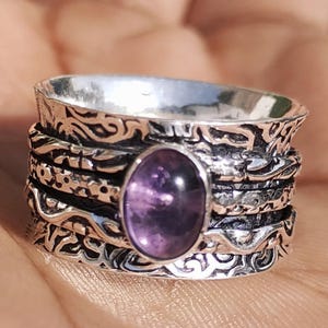 May include: A silver-colored spinner ring with an oval, purple gemstone. The ring has intricate, etched patterns and a textured design. The gemstone is set in the center of the ring.