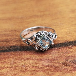 Könnte beinhalten: Ein silberner Ring mit einem quadratischen, hellblauen Edelstein in der Mitte. Der Ring hat ein verziertes Design mit Wirbeln und kleinen, runden Verzierungen. Das Band ist glatt und poliert, auf einer Holzoberfläche.