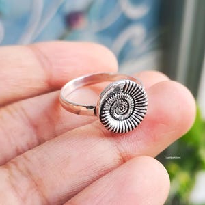 Pu&ograve; includere: Un anello d'argento con un design a spirale dettagliato. L'anello presenta un elemento circolare strutturato con un motivo a spirale prominente, montato su una fascia sottile e lucida. L'anello &egrave; mostrato sul dito di una persona.