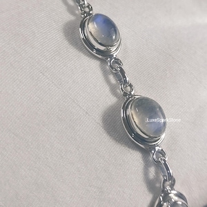 Könnte beinhalten: Ein silbernes Armband mit ovalen Mondstein-Edelsteinen. Die Mondsteine haben ein milchiges, schillerndes Aussehen und sind in eine silberne Lünette gefasst. Das Armband ist mit kleinen silbernen Kettengliedern verbunden. Der Text "LuxeSparkStone" ist sichtbar.