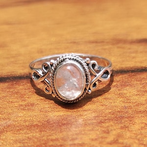 Pu&ograve; includere: Un anello d'argento con una gemma ovale di colore chiaro. La gemma &egrave; incastonata in una lunetta dettagliata con un motivo a corda intrecciata. L'anello presenta volute decorative sui lati, creando un aspetto elegante e ornato.