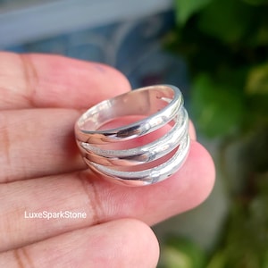 Pode incluir: Um anel de prata polida com um design de múltiplas bandas, apresentando três aros de metal distintos, lisos e arredondados, conectados na parte de trás. A superfície do metal é reflexiva e brilhante, com um acabamento metálico claro.