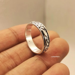 Könnte beinhalten: Ein silberner Ring mit floralem Design. Der Ring hat ein Band mit detaillierten Schnitzereien von Blumen und Blättern. Der Ring wird in einer Hand gehalten, was sein aufwendiges Design und seine polierte Oberfläche hervorhebt. Der Ring ist ein Schmuckstück.