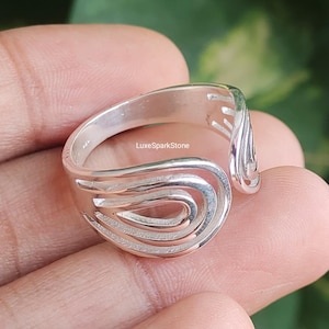 Może przedstawiać: Srebrny pierścionek o otwartym wzorze z szeregiem zakrzywionych linii. Pierścionek ma gładkie, wypolerowane wykończenie i jest pokazany na palcu. Projekt przypomina płynącą wodę lub stylizowaną falę.