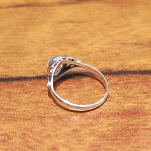 Pu&ograve; includere: Un anello d'argento con una pietra di forma ovale e di colore chiaro. L'anello presenta un design dettagliato e ornato attorno alla pietra e lungo il gambo. Appoggiato su una calda superficie di legno.