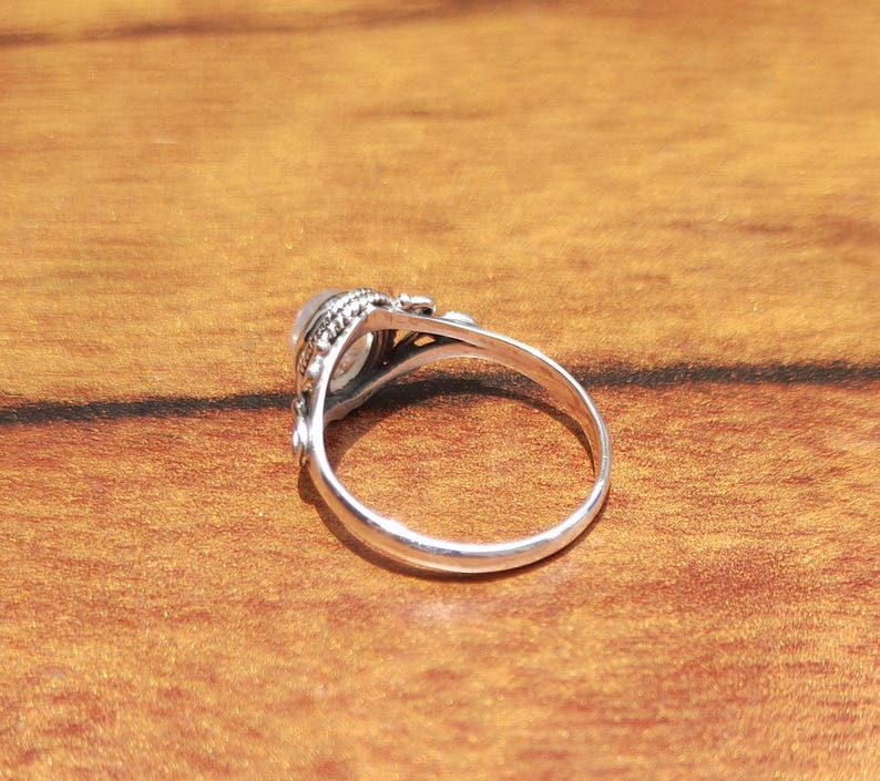 Pu&ograve; includere: Un anello d'argento con una pietra di forma ovale e di colore chiaro. L'anello presenta un design dettagliato e ornato attorno alla pietra e lungo il gambo. Appoggiato su una calda superficie di legno.