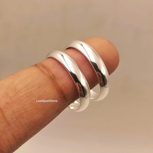 Könnte beinhalten: Zwei polierte silberfarbene Ringe mit einem glatten, abgerundeten Bandprofil. Die Ringe haben eine hochglänzende, reflektierende Oberfläche und ein schlichtes, schmuckloses Design. Der Text LuxeSparkStone ist auf dem unteren Ringband sichtbar.