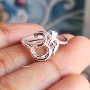 Op de afbeelding: Een zilverkleurige ring met het Om-symbool. De ring wordt close-up getoond, waarbij het ingewikkelde ontwerp en de gepolijste afwerking worden benadrukt. De ring wordt in een hand gehouden, met een vage achtergrond.