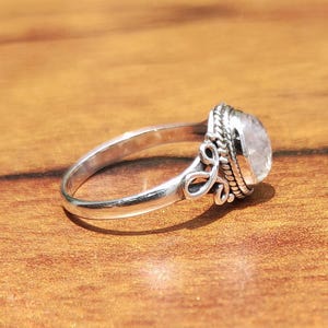 Pu&ograve; includere: Un anello d'argento con una gemma ovale e traslucida. L'anello presenta un design ornato attorno alla gemma e sulla fascia. L'anello &egrave; appoggiato su una superficie di legno.