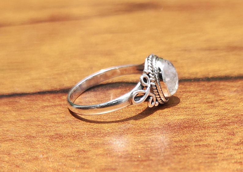 Pu&ograve; includere: Un anello d'argento con una gemma ovale e traslucida. L'anello presenta un design ornato attorno alla gemma e sulla fascia. L'anello &egrave; appoggiato su una superficie di legno.