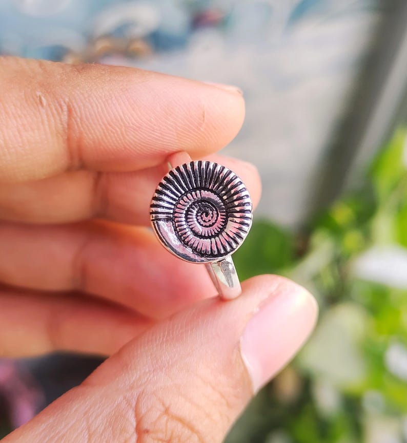 Pu&ograve; includere: Un anello d'argento con un design a spirale dettagliato. L'anello presenta un emblema circolare con linee concentriche, simile a un fossile. L'anello &egrave; tenuto tra le dita su uno sfondo sfocato verde e blu.