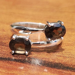May include: A silver ring with two oval, dark brown gemstones. The ring's band splits to hold each stone, creating a unique design. The gemstones are set in a prong setting, and the ring is displayed on a wooden surface.