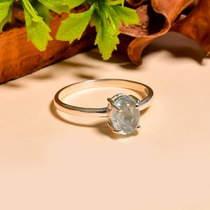 Könnte beinhalten: Ein silberner Ring mit einem hellblauen, ovalen Edelstein. Der Ring liegt auf einer hellbeigen Oberfläche, mit grünen Blättern und braunem Holz im Hintergrund. Der Edelstein ist der Mittelpunkt des Rings.
