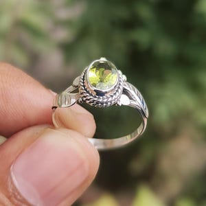 Peut inclure: Une bague en argent ornée d'une pierre précieuse ovale vert clair. La pierre est sertie dans une lunette détaillée en forme de corde. La bague présente des accents décoratifs en forme de feuilles sur les côtés et un anneau poli.