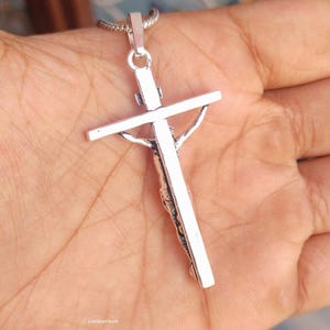 Puede incluir: Un colgante de crucifijo plateado con una representaci&oacute;n detallada de Jes&uacute;s en la cruz. El colgante cuelga de una delicada cadena. La imagen es un primer plano, que destaca los intrincados detalles de la joyer&iacute;a religiosa.
