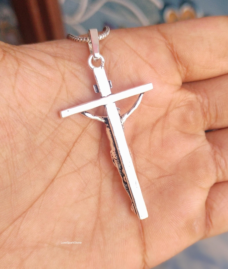 Puede incluir: Un colgante de crucifijo plateado con una representaci&oacute;n detallada de Jes&uacute;s en la cruz. El colgante cuelga de una delicada cadena. La imagen es un primer plano, que destaca los intrincados detalles de la joyer&iacute;a religiosa.