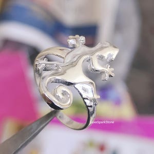 May include: A silver-colored ring featuring a detailed lizard design. The lizard is depicted with intricate scales and a curled tail. The ring is held by a pair of tweezers, with the LuxeSparkStone logo visible in the background.