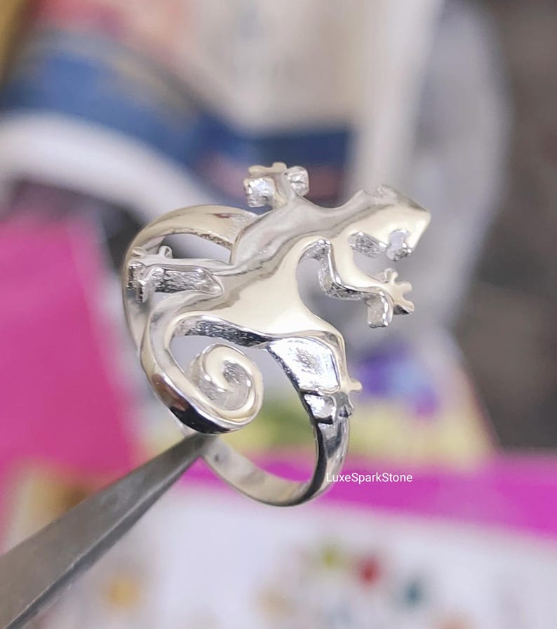 May include: A silver-colored ring featuring a detailed lizard design. The lizard is depicted with intricate scales and a curled tail. The ring is held by a pair of tweezers, with the LuxeSparkStone logo visible in the background.