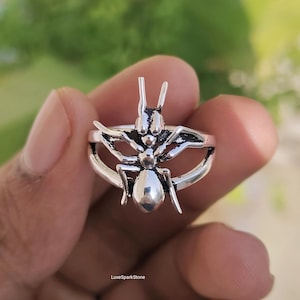 Könnte beinhalten: Ein silberner Ring mit einem Ameisen-Design. Der Ring hat eine detaillierte Insektenfigur mit Antennen, einem segmentierten Körper und gebogenen Flügeln. Der Ring wird in einer Hand vor einem verschwommenen grünen Hintergrund gehalten. Der Text "LuxeSparkStone" ist sichtbar.