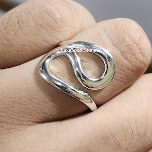 May include: A silver ring with an abstract design. The ring has a continuous, flowing shape with a loop and a curved band. The ring is worn on a finger, highlighting its modern and minimalist style. The text "LuxeSparkStone" is visible.