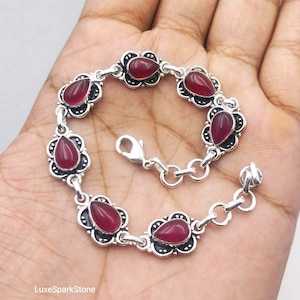 Könnte beinhalten: Ein Silberarmband mit sechs tropfenförmigen, dunkelroten Edelsteinen, die in kunstvoll verzierten, gewellten Metallfassungen sitzen. Die Glieder sind durch kleine Silberringe verbunden und das Stück verfügt über einen Karabinerverschluss.