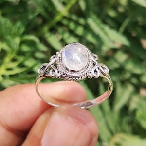 Könnte beinhalten: Ein silberner Ring mit einem ovalen, schillernden Mondstein. Der Ring hat ein verziertes Design mit Wirbeldetails um den Stein und entlang des Bandes. Der Mondstein hat ein milchiges, opaleszierendes Aussehen.