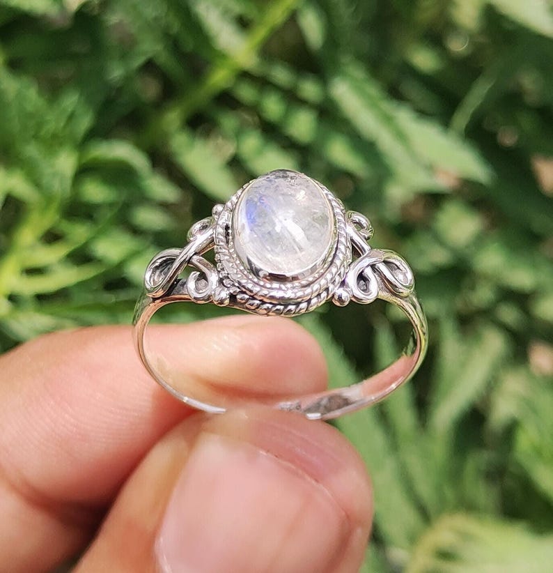 Pu&ograve; includere: Un anello d'argento con una pietra di luna ovale e iridescente. L'anello presenta un design ornato con dettagli a spirale attorno alla pietra e lungo il gambo. La pietra di luna ha un aspetto lattiginoso e opalescente.
