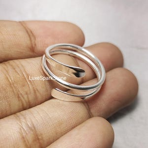 Könnte beinhalten: Ein silberfarbener Wickelring mit Spiraldesign. Der Ring hat eine glatte, polierte Oberfläche und wickelt sich mit einem offenen, verstellbaren Design um den Finger. Der Ring wird auf einer Hand gezeigt.