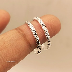 Könnte beinhalten: Zwei silberne Ringe mit einem sich wiederholenden geprägten Muster aus kleinen Rautenformen und floralen Motiven. Das Metall hat eine polierte Oberfläche mit abgedunkelten vertieften Bereichen, um die Details hervorzuheben. Der Text LuxeSparkStone ist sichtbar.