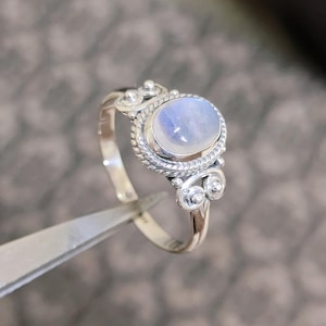 Könnte beinhalten: Silberring mit einem ovalen, schillernden Mondstein. Der Stein ist in einer detaillierten Lünette mit einem gedrehten Seildesign gefasst. Der Ring hat dekorative Wirbel an den Seiten.
