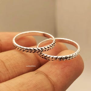 Könnte beinhalten: Zwei silberne Ringe mit einem sich wiederholenden Blattmuster um das Band. Die Ringe werden auf dem Finger einer Person gezeigt. Die Ringe sind hellsilberfarben und haben ein zartes, detailliertes Design. Die Ringe sind wahrscheinlich für den täglichen Gebrauch.