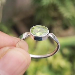 May include: A silver ring with a light green, oval gemstone. The gemstone is set in a bezel setting, and the band has a simple design. The ring is displayed against a blurred green background.
