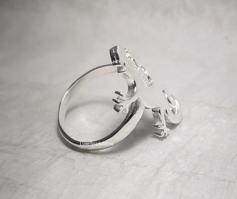 May include: A silver-colored ring featuring a lizard design. The lizard is detailed with visible claws and a textured body. The ring band is smooth and polished. The ring is shown on a neutral background.