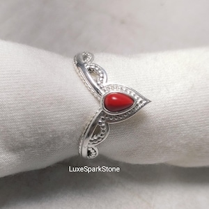 Può includere: Un anello in argento con una pietra rossa a forma di goccia. L'anello presenta un design dettagliato e ornato con una fascia strutturata e dettagli decorativi. L'anello è mostrato su uno sfondo bianco. Il testo "LuxeSparkStone" è visibile.