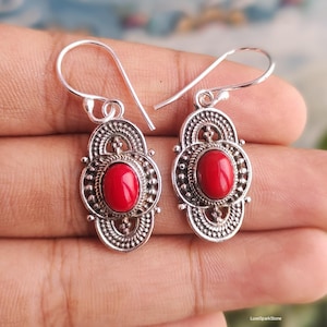 May include: A pair of silver-toned earrings with a red oval stone center. The earrings have a decorative filigree design around the stone and a hook closure. The earrings are displayed on a hand.