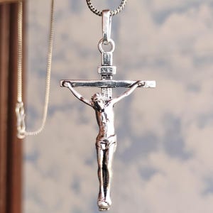 Puede incluir: Un colgante de crucifijo plateado con una detallada representaci&oacute;n de Jesucristo en la cruz. El colgante cuelga de una delicada cadena plateada. El fondo es un cielo suave y borroso.