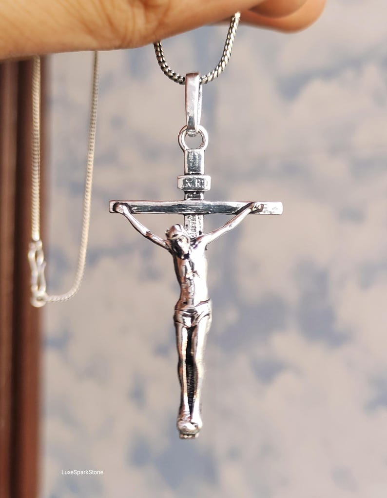 Puede incluir: Un colgante de crucifijo plateado con una detallada representaci&oacute;n de Jesucristo en la cruz. El colgante cuelga de una delicada cadena plateada. El fondo es un cielo suave y borroso.