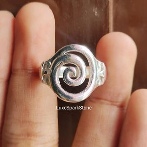May include: A silver ring with a spiral design. The ring features a central swirl, with a band that widens at the sides. The ring is held between fingers, with the LuxeSparkStone logo visible.