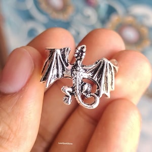 Könnte beinhalten: Ein silberner Drachenring mit detaillierten Flügeln, Körper und Schwanz. Der Drache ist in einer Flugpose dargestellt, mit weit ausgebreiteten Flügeln. Der Ring wird in einer Hand gehalten und zeigt das aufwendige Design und die Handwerkskunst des Fantasy-Schmucks.